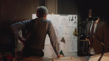 Male mature tailor works in luxury designer atelier or tailoring studio. He looks at board with sketches of future clothes and thinks. Mannequin with tailored suit. Fashion and hand craft concept.