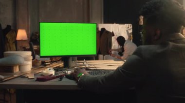 African American tailor or fashion designer creates and developes clothes on computer with green screen in luxury atelier or tailoring workshop. Man sits on workplace and types on keyboard. Chromakey.