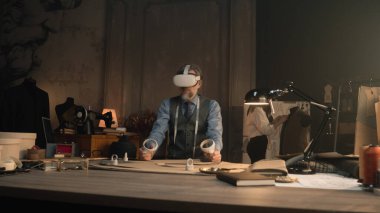 Mature tailor wearing VR headset with wireless controllers, sits at the table in luxury designer atelier, gesturing and creates design in virtual reality. Concept of technologies of future in business