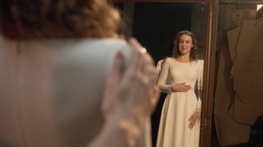 Female tailor zips up wedding dress on woman in front of the mirror. Happy bride on fitting wedding dress in luxury designer atelier or wedding salon. Concept of fashion, handmade and couturier.