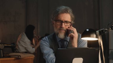 Mature tailor talks with client by phone, takes order online using laptop, discusses colors and textile of tailored suit. He works in stylish luxury designer atelier. Concept of fashion and handmade.