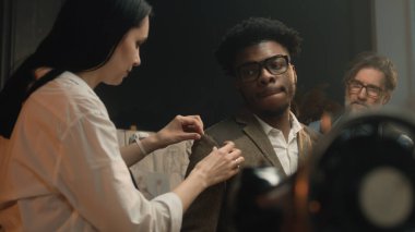 Female tailor measures tailored suit, discusses with client size and fabric. African American client on fitting wedding business jacket in luxury atelier or tailoring workshop. Fashion concept.