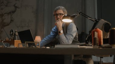 Mature tailor in black framed glasses works on new clothes collection. Man draws modern design of tailorer suit on laptop or tablet computer. Concept of fashion and craftsman job at workshop.