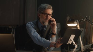 Mature tailor talking by phone and working on design of suit or new clothes collection using tablet computer and laptop. He works in stylish luxury designer atelier. Concept of fashion and handmade.