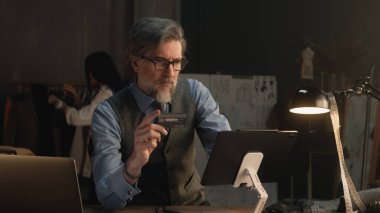 Tailor using tablet, transferring money on bank card, paying for service online during work in luxury atelier. Female tailor works on elegant suit on background. Fashion and entrepreneurship concept.