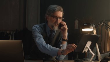 Mature tailor talking by phone and working on design of suit or new clothes collection using tablet computer and laptop. He works in stylish luxury designer atelier. Concept of fashion and handmade.