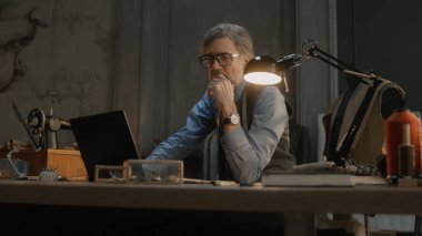 Mature tailor in black framed glasses works on new clothes collection. Man draws modern design of tailorer suit on laptop or tablet computer. Concept of fashion and craftsman job at workshop.