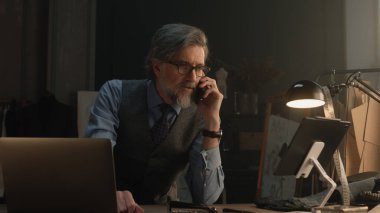 Mature tailor talking by phone and working on design of suit or new clothes collection using tablet computer and laptop. He works in stylish luxury designer atelier. Concept of fashion and handmade.