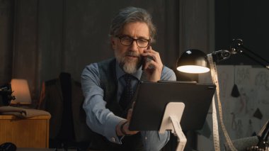 Mature tailor talking by phone and working on design of suit or new clothes collection using tablet computer and laptop. He works in stylish luxury designer atelier. Concept of fashion and handmade.