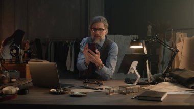 Mature tailor in black framed glasses chatting with client by the phone, taking the order in luxury designer atelier. Woman works with wedding suit on background. Concept of fashion and hand craft.
