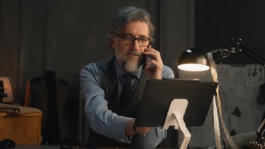 Mature tailor talking by phone and working on design of suit or new clothes collection using tablet computer and laptop. He works in stylish luxury designer atelier. Concept of fashion and handmade.