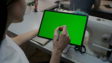 Wear designer sits at sewing machine, keeps stylus in hand, watches at tablet and touches screen. Chroma key displayed on horizontal placed device. Atelier workshop. Concept of fashion. Close up shot.