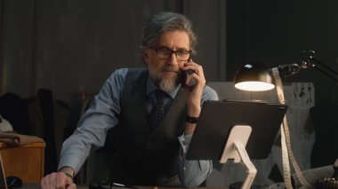 Mature tailor talking by phone and working on design of suit or new clothes collection using tablet computer and laptop. He works in stylish luxury designer atelier. Concept of fashion and handmade.