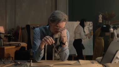 Tired male tailor sits at table with tailoring tools, patterns, tablet and laptop. He takes off glasses and thinks. Female tailor works on dress on background. Fashion, style and hand craft concept.