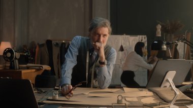Tired male tailor sits at table with tailoring tools, patterns, tablet and laptop. He takes off glasses and thinks. Female tailor works on dress on background. Fashion, style and hand craft concept.
