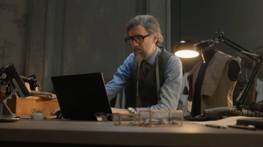 Mature tailor in black framed glasses works on new clothes collection. Man draws modern design of tailorer suit on laptop or tablet computer. Concept of fashion and craftsman job at workshop.