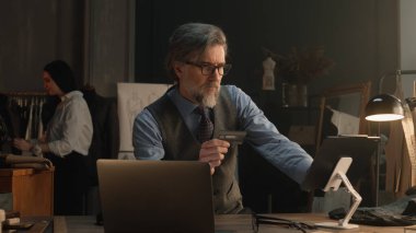 Tailor using tablet, transferring money on bank card, paying for service online during work in luxury atelier. Female tailor works on elegant suit on background. Fashion and entrepreneurship concept.