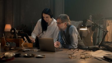 Tailors choosing design of tailored clothes collection on laptop and digital tablet computer. Colleagues working at modern workshop, talking about styles of garment, sewing type and color of cloth.