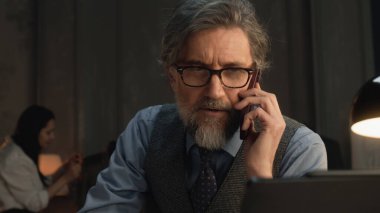 Mature tailor talking by phone and working on design of suit or new clothes collection using digital tablet and laptop. He works in stylish luxury designer atelier. Concept of fashion and handmade.