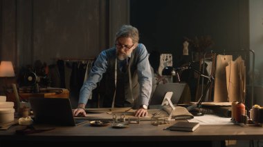 Mature man creates clothes design for new wear collection using tablet and laptop. Tailor in glasses looks at camera, stands near table with tailoring tools. Concept of fashion and hand craft.