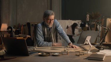 Tired male tailor sits at table with tailoring tools, patterns, tablet and laptop. He takes off glasses and thinks. Female tailor works on dress on background. Fashion, style and hand craft concept.