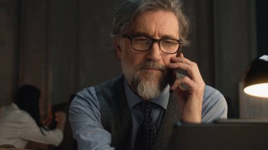 Mature tailor talking by phone and working on design of suit or new clothes collection using digital tablet and laptop. He works in stylish luxury designer atelier. Concept of fashion and handmade.