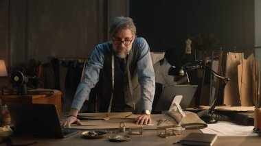 Mature man creates clothes design for new wear collection using tablet and laptop. Tailor in glasses looks at camera, stands near table with tailoring tools. Concept of fashion and hand craft.