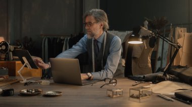 Mature couturier in black glasses works on new wear collection in atelier. Man draws modern design of tailored suit on laptop or tablet computer. Concept of fashion and craftsman job. Slow motion