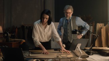 Tailors working with patterns for future clothes in luxury designer atelier. Mature man showing time on his clocks and proposes to take a break. Concept of coworking, fashion and hand craft.