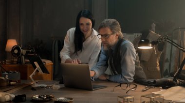 Tailors choosing design of tailored clothes collection on laptop and digital tablet computer. Colleagues working at modern workshop, talking about styles of garment, sewing type and color of cloth.