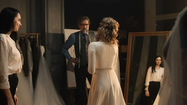 Female tailor zips up wedding dress on woman in front of mirror. Bride on fitting wedding dress in luxury designer atelier or wedding salon. Concept of fashion, handmade and couturier. Slow motion.