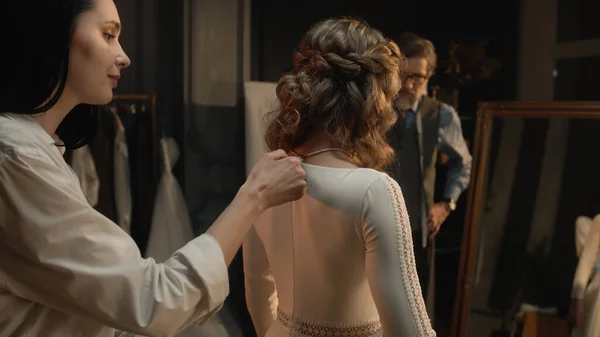 Cinematic shot: female tailor zips up wedding dress on woman in front of mirror. Bride on fitting wedding dress in luxury designer atelier or wedding salon. Concept of fashion, handmade and couturier.
