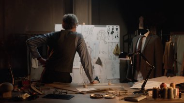 Male mature tailor works in luxury designer atelier or tailoring studio. He looks at board with sketches of future clothes and thinks. Mannequin with tailored suit. Fashion and hand craft concept.