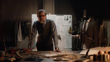 Male mature tailor works in luxury designer atelier or tailoring studio, looks at camera. Table with threads, fabric and tools. Tailored suits and board with sketches on background. Fashion concept.