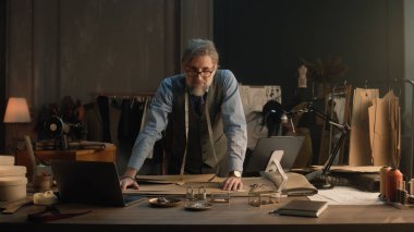 Mature man creates clothes design for new wear collection using tablet and laptop. Tailor in glasses looks at camera, stands near table with tailoring tools. Concept of fashion and hand craft.