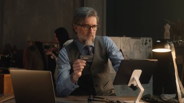 Tailor using tablet, transferring money on bank card, paying for service online during work in luxury atelier. Female tailor works on elegant suit on background. Fashion and entrepreneurship concept.