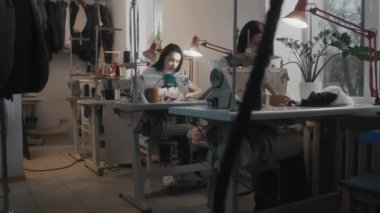 Female seamstresses sitting at sewing machines and working on custom apparel. Measure tapes and laptop with sketch of wedding suit at the table. Atelier or sewing workshop. Concept of fashion.