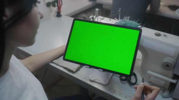 女裁缝或女裁缝师坐在缝纫机前 手握手写笔 看着平板电脑 在水平放置装置上显示绿色屏幕 Atelier车间 时尚的概念 近距离拍摄 — 图库视频影像