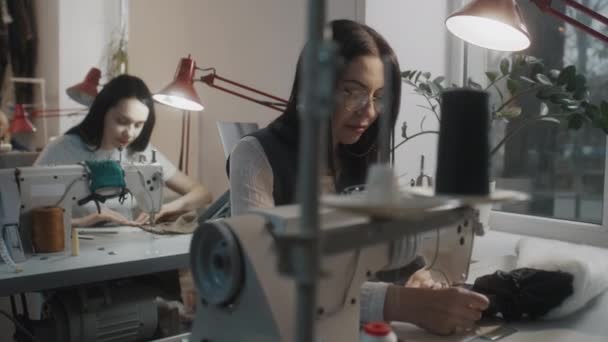 戴眼镜的女裁缝坐在缝纫机前 缝制定制服装 西默斯的同事们在桌边工作 背景是笔记本电脑 大堂或缝纫车间 时尚的概念 — 图库视频影像