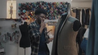 African American fashion designer stands near the mannequin in unfinished tailored wedding or business suit with digital tablet computer and thinks. Seamstresses working on background in atelier.