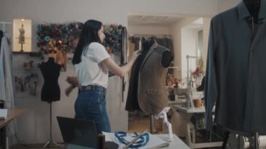 Dressmaker keeps in hand tablet, zoomes sketch and touches wedding suit. Seamstresses sits and works at sewing machines. Threads rack and mannequin at background. Atelier workshop. Fashion concept.