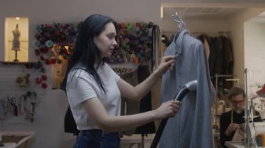 Seamstress irons new tailored shirt with electric hand steam generator in atelier workshop. Male tailor sews clothes on background. Concept of small business, tailoring industry and fashion.