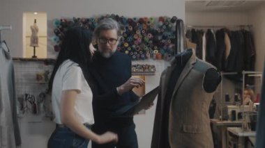 Male mature fashion designer shows sketch of future jacket to seamstress on laptop in atelier workshop and discusses tailoring process. Mannequin in unfinished suit. Fashion and hand craft concept.