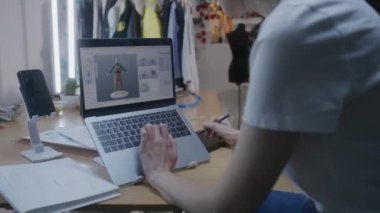 Female fashion designer draws future clothes in 3D modeling software on laptop using digital tablet computer and stylus. She works in atelier workshop. Concept of fashion and technologies in business.