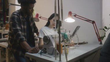 African American tailor with seamstress discussing sketch of wedding suit displayed on tablet. Female dressmaker sits at sewing machine and touches device. Atelier or sewing workshop. Fashion concept.