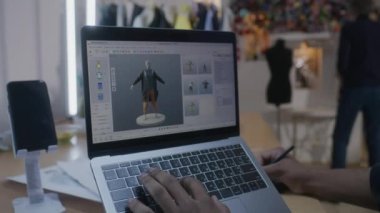 African American fashion designer creates clothes in 3D modeling software on laptop using digital tablet and stylus. He works in atelier workshop. Concept of fashion and technologies in business.