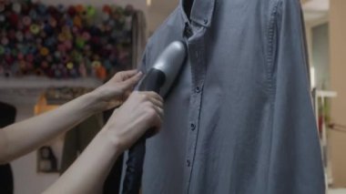 Close-up shot of woman irons tailored shirt with electric handheld steamer in atelier workshop. Male tailor sews clothes on background. Concept of small business, tailoring industry and fashion.