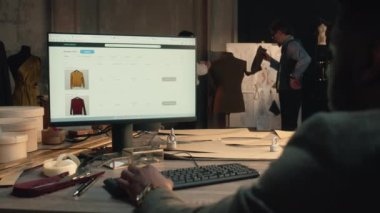 African American man browsing personal clothing shop on PC or watching orders statistic. Businessman sits at the table at luxury designer atelier and works with range on website. Concept of fashion.