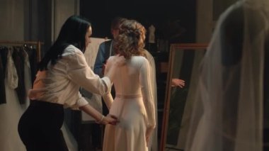 Female tailor zips up wedding dress on woman in front of mirror. Bride on fitting wedding dress in luxury designer atelier or wedding salon. Concept of fashion, handmade and couturier. Slow motion.