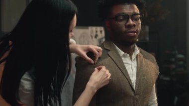 Female tailor measures tailored suit, discusses with client size and fabric. African American client on fitting wedding business jacket in luxury atelier or tailoring workshop. Fashion concept.